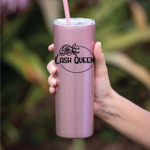 Pode incluir: Um copo de aço inoxidável cor ouro rosa com um design preto que diz "LASH QUEEN" com um gráfico de rosa dentro de um círculo. O copo tem um canudo rosa.