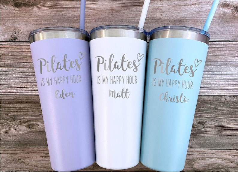 May include: Three stainless steel tumblers with lids and straws. The tumblers are lavender, white, and light blue. Each tumbler has the text "Pilates is my happy hour" with a heart symbol and a name below: Eden, Matt, and Christa.