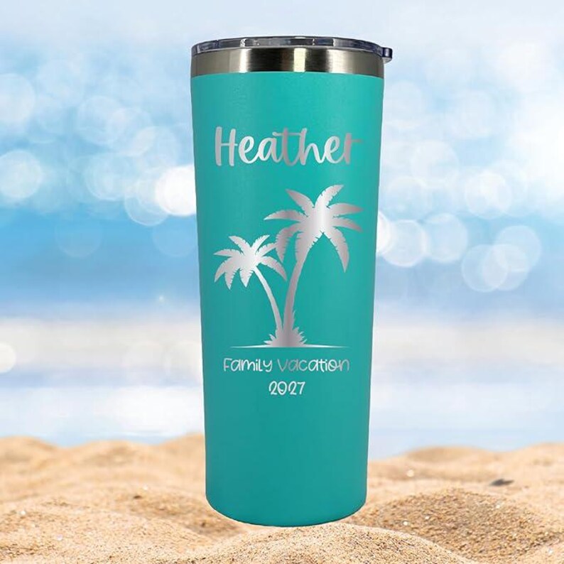 May include: A turquoise stainless steel tumbler with a silver lid. The tumbler has a silver palm tree design and the text "Heather Family Vacation 2027" printed on it.