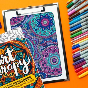 May include: A coloring book with the title "The Art Therapy Adult Coloring Book" and a colorful mandala design on the cover. The book is on a clipboard with a variety of colored markers and pencils on the right side of the image.