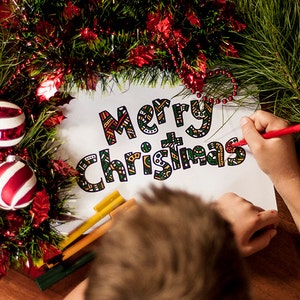 May include: A child colors in a "Merry Christmas" sign with red, green, and yellow markers. The sign is surrounded by a green garland with red and white ornaments.