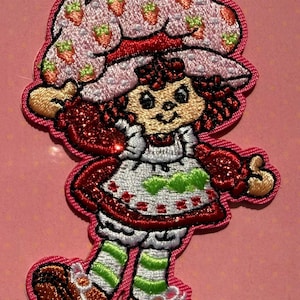 May include: An embroidered iron-on patch featuring the character Strawberry Shortcake. The character wears a large pink hat with strawberry patterns, a white dress with red accents, green and white striped leggings, and brown shoes.