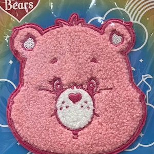 May include: A pink Care Bear patch with a textured surface. The bear's face features white accents around the eyes and mouth, and a heart-shaped nose. The Care Bears logo is visible in the upper left corner.