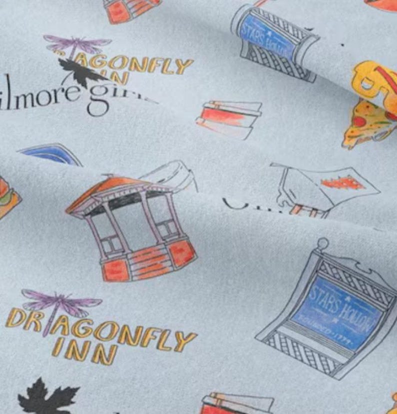 May include: A light blue fabric with a repeating pattern of various illustrations. The illustrations include a dragonfly, a gazebo, a pizza slice, a sign that says "Stars Hollow", and the text "Dragonfly Inn".