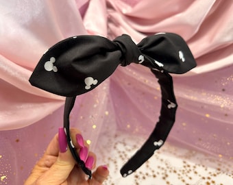 May include: A black fabric headband featuring a large, tied bow at the top. The headband is decorated with a repeating white pattern of small, circular mouse-ear silhouettes distributed across the black material of the bow and the band.