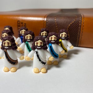 May include: A group of small figurines depicting figures with brown hair and beards, wearing white robes and colorful sashes. They stand in front of a brown leather-bound book. The figurines have smiling faces and are on a white surface.