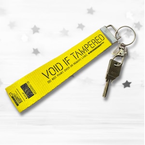 K&ouml;nnte beinhalten: Ein gelbes Schl&uuml;sselband mit dem Text "VOID IF TAMPERED" in Schwarz. Das Band ist an einem silbernen Schl&uuml;sselring und einem Schl&uuml;ssel befestigt. Der Hintergrund ist wei&szlig; mit grauen Sternformen.