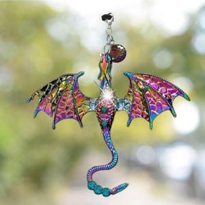 May include: A colorful dragon ornament with iridescent wings and a long, curved tail. The dragon features a jewel-like body with teal and purple accents. It hangs from a silver chain and a black clip.