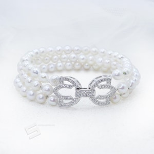 May include: A three-strand pearl bracelet with a silver bow-shaped clasp. The pearls are white and round, and the clasp is encrusted with small, clear gemstones. The bracelet is displayed on a white surface.