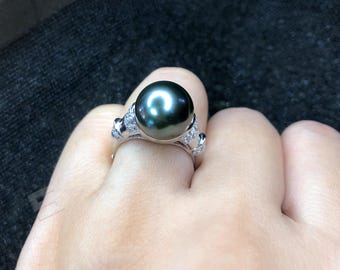 Könnte beinhalten: Ein silberner Ring mit einer großen, dunkelgrauen Perle. Der Ring ist mit kleinen, klaren Edelsteinen um die Perle und an den dekorativen Elementen des Rings verziert. Der Ring wird an einem Finger getragen.