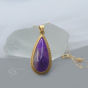 May include: A gold-colored necklace with a teardrop-shaped pendant. The pendant features a polished, purple gemstone with a gold rope-like border. The necklace is displayed on a light gray surface.