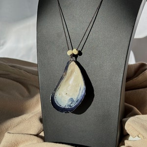 Può includere: Una collana con un grande ciondolo a forma di conchiglia di mare iridescente. Il ciondolo è di colore beige chiaro con un bordo blu scuro. La collana è realizzata con un cordoncino marrone scuro con due piccole perle marroni chiare.