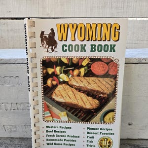 May include: A vintage Wyoming Cookbook with a spiral binding. The cover features a colorful illustration of a cowboy riding a horse and a close-up of grilled steaks and vegetables. The text on the cover reads "Wyoming Cookbook" and lists various recipes, including Western, Beef, Fresh Garden Produce, Homemade Pastries, Wild Game, Pioneer, Dessert Favorites, Fruit, Fish, and Trivia. The book is part of the "Cooking Across America Series."