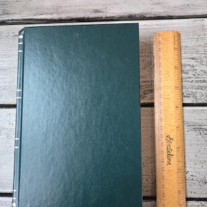 May include: A dark green hardback book with white stitching along the spine. The book is lying flat on a wooden surface next to a wooden ruler.