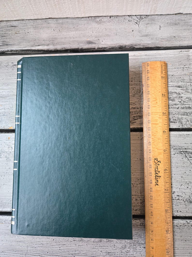 May include: A dark green hardback book with white stitching along the spine. The book is lying flat on a wooden surface next to a wooden ruler.