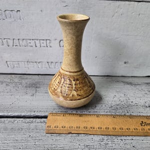 May include: A small, tan ceramic vase with a bulbous base and a slender neck. The base features a brown, textured pattern. The vase is approximately 5 inches tall, as measured by a wooden ruler.