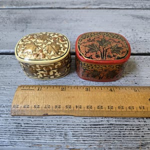 May include: Two small, oval-shaped decorative boxes. One is cream-colored with a brown leaf design, and the other is red with a black floral pattern. A wooden ruler with inch markings is placed below the boxes.