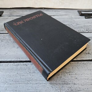 May include: A hardcover book titled "The Apostle" with a black cover and red lettering. The spine and edges are brown. The book is resting on a weathered, light gray wooden surface.
