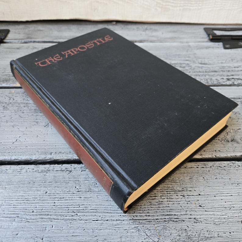 May include: A hardcover book titled "The Apostle" with a black cover and red lettering. The spine and edges are brown. The book is resting on a weathered, light gray wooden surface.