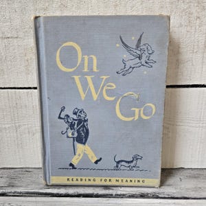 May include: A gray hardcover book titled On We Go with yellow lettering. The cover features a black illustration of a person walking with a pack and a dog, a winged dog above, and a small dog below. The text Reading for Meaning is at the bottom.