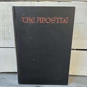 May include: A hardcover book with a black cover. The title "THE APOSTLE" is printed in red, stylized lettering across the top of the book. The book is slightly angled, revealing the spine and edges.