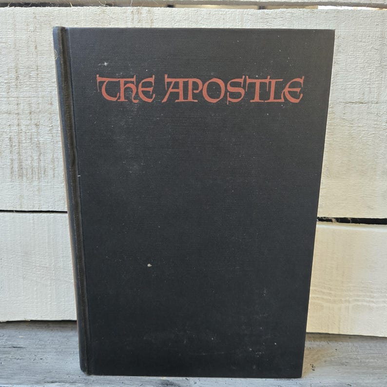 May include: A hardcover book with a black cover. The title "THE APOSTLE" is printed in red, stylized lettering across the top of the book. The book is slightly angled, revealing the spine and edges.