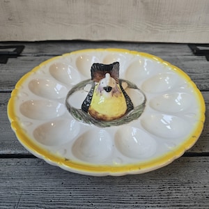 May include: A round ceramic egg serving platter featuring a central three-dimensional figure of a hen sitting in a nest. The platter has twelve oval indentations for eggs and a scalloped yellow rim. The hen is painted in yellow, black, and brown.