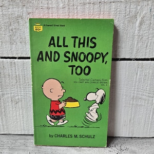 May include: A vintage paperback book titled "ALL THIS AND SNOOPY, TOO" by Charles M. Schulz. The cover features Charlie Brown holding a food bowl for Snoopy. The book has a green cover with white and black text and illustrations.
