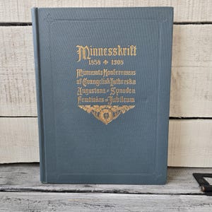May include: A vintage book with a blue-gray cover. The cover features gold lettering that reads "Minnesskrift 1858-1908" and other text. A decorative gold emblem is at the bottom. The book is closed and appears to be in good condition.