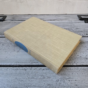 May include: A hardcover book with a beige linen cover. A blue oval label is on the spine. The book is on a weathered, gray and white wooden surface.