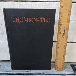 May include: A black hardcover book titled "THE APOSTLE" in red lettering. A wooden ruler is positioned next to the book. The book appears to be vintage, with a simple, classic design. The ruler is marked with inch measurements.