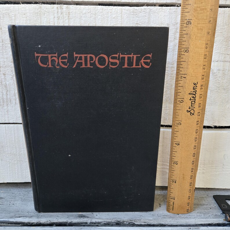 May include: A black hardcover book titled "THE APOSTLE" in red lettering. A wooden ruler is positioned next to the book. The book appears to be vintage, with a simple, classic design. The ruler is marked with inch measurements.