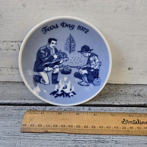 May include: A blue and white decorative plate with the text "Fars Dag 1972". The plate features a scene of two figures, one kneeling and the other seated, cooking over a campfire. A ruler is placed next to the plate.