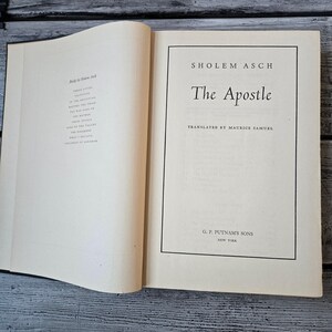 May include: An open book with the title "The Apostle" by Sholem Asch. The book's cover is cream-colored with black text. The publisher's name, G. P. Putnam's Sons, is also visible. The book is on a wooden surface.
