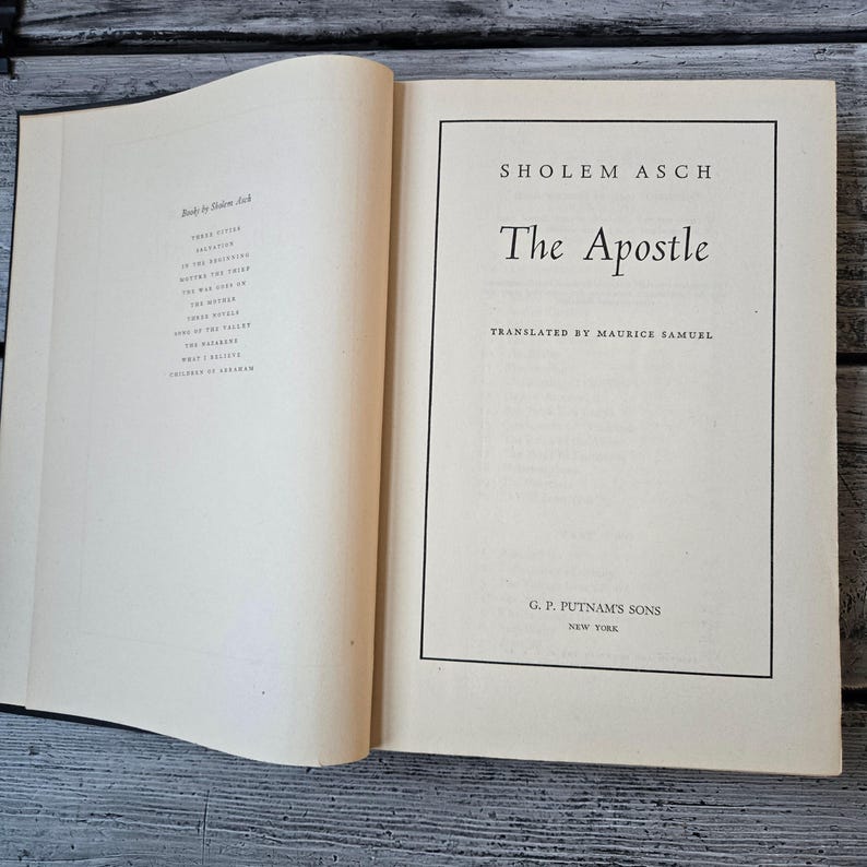 May include: An open book with the title "The Apostle" by Sholem Asch. The book's cover is cream-colored with black text. The publisher's name, G. P. Putnam's Sons, is also visible. The book is on a wooden surface.