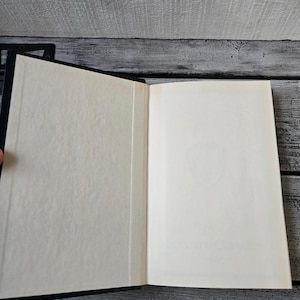 May include: A black hardback book with a blank white interior. The book is open and lying flat on a wooden surface.