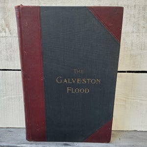 May include: An antique book titled "The Galveston Flood." The book has a dark gray cover with burgundy accents on the spine and corners. The title is in gold lettering.