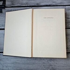 May include: An open book with off-white pages. The right page displays the title "THE APOSTLE" in a centered, serif font. The book's spine is visible, and the cover is black. The book rests on a weathered, gray wooden surface.