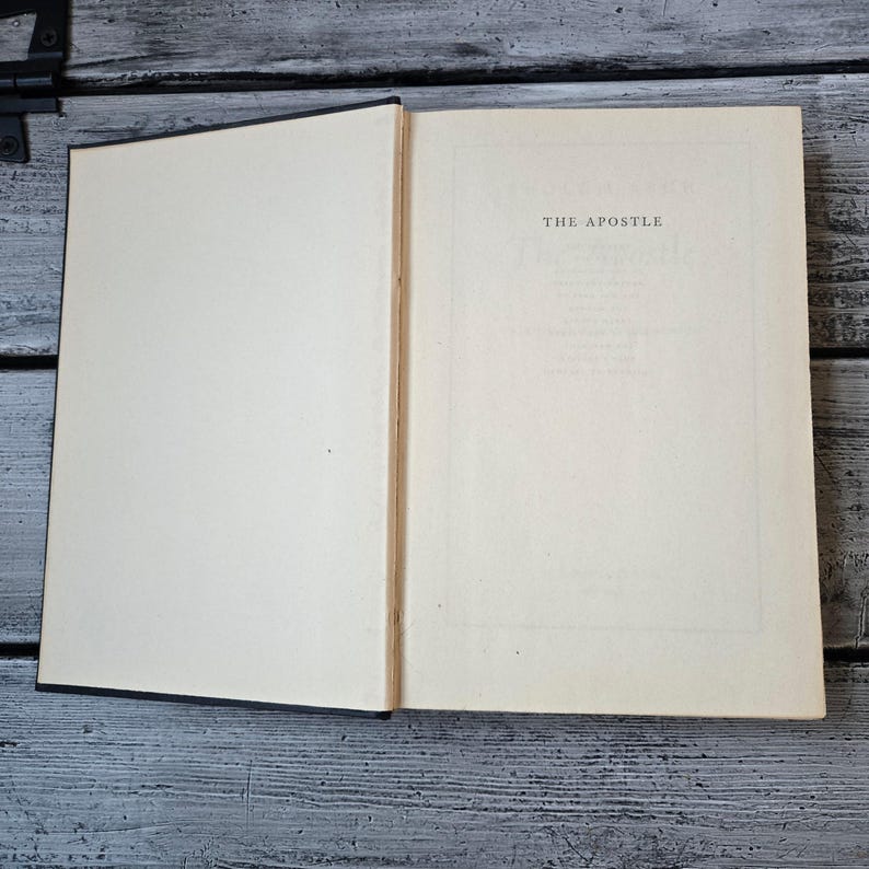 May include: An open book with off-white pages. The right page displays the title "THE APOSTLE" in a centered, serif font. The book's spine is visible, and the cover is black. The book rests on a weathered, gray wooden surface.