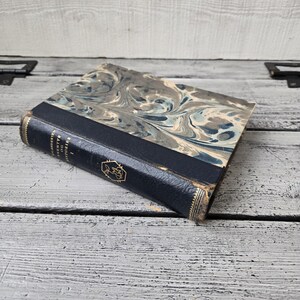 May include: An antique book with a marbled cover in shades of blue, gray, and white. The spine is dark blue with gold lettering and a decorative emblem. The book is resting on a weathered, light-colored wooden surface.