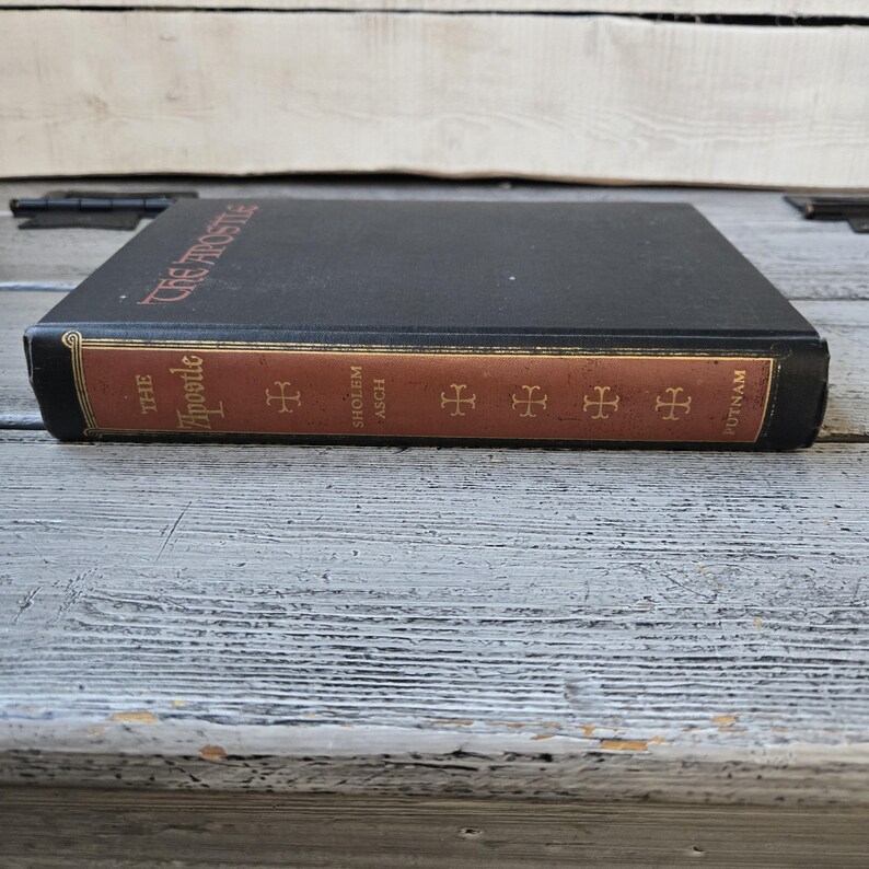 May include: A hardcover book titled "The Apostle" with a black cover and a brown spine. The spine features the title, author, and publisher. The book is resting on a weathered, light-colored wooden surface.