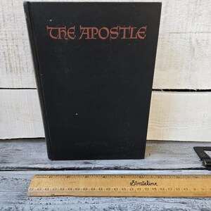 May include: A black book titled "THE APOSTLE" with red lettering. A wooden ruler with inch markings is at the bottom of the image. The book is standing upright on a white and gray surface.