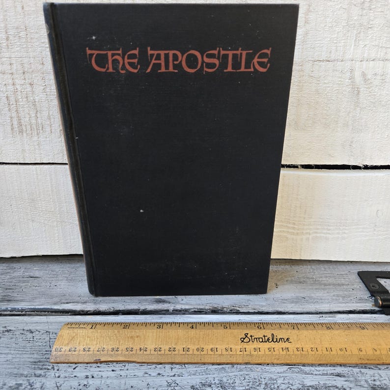 May include: A black book titled "THE APOSTLE" with red lettering. A wooden ruler with inch markings is at the bottom of the image. The book is standing upright on a white and gray surface.