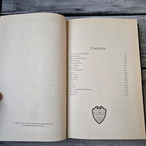 May include: A book titled "The American Scholar" by Ralph Waldo Emerson. The book contains a table of contents with essays such as "Self-reliance", "Compensation", "Friendship", and "Heroism".
