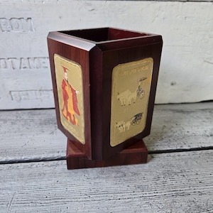 May include: A dark wood pen holder with gold rectangular panels featuring illustrations of figures and vehicles. The pen holder has a square top and a smaller square base.
