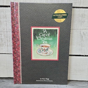 May include: A hardcover book titled "A Cup of Christmas Tea" with a watercolor illustration of a teacup and saucer. The cover is dark gray with white polka dots and a red floral spine. An "Autographed Copy" sticker is on the cover.
