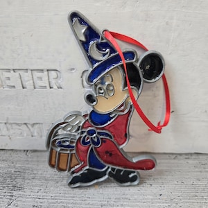 May include: A colorful Mickey Mouse ornament in the Sorcerer's Apprentice attire. The ornament features a red, blue, and silver color scheme with a red ribbon for hanging. The design is a classic Disney character.