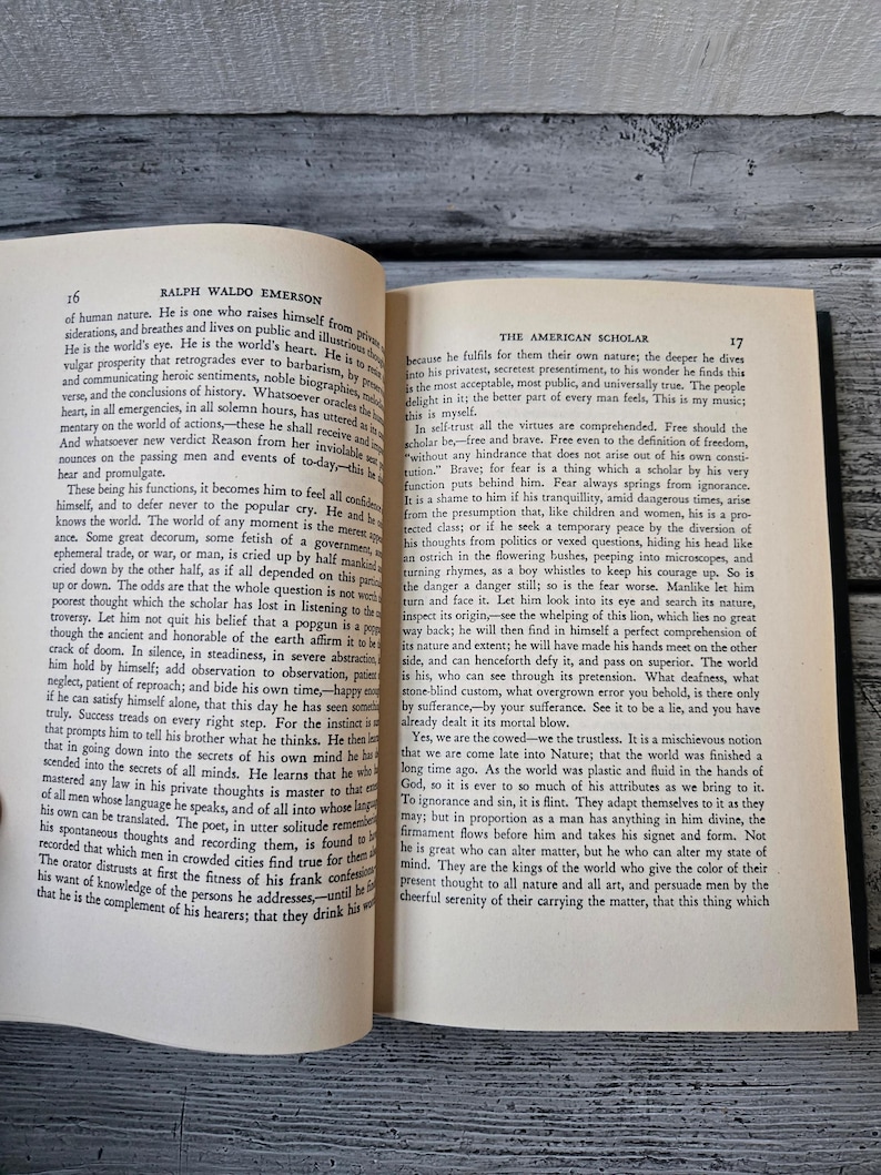 May include: An open book with a tan cover and black text. The book is titled "The American Scholar" by Ralph Waldo Emerson. The text on the page discusses the importance of self-trust and bravery for a scholar.