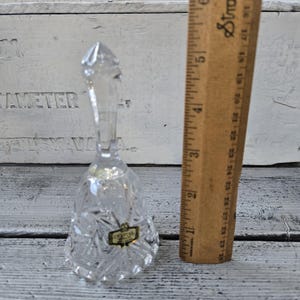 May include: A clear crystal bell with a faceted design and a long handle. A wooden ruler is next to the bell. The bell has a gold label that reads "Crystal Hand Cut".