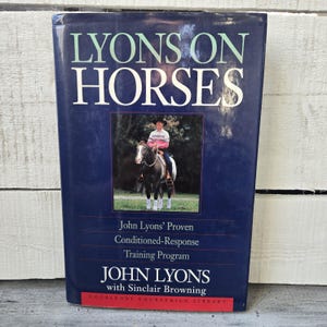 May include: A hardcover book titled "LYONS ON HORSES" with a dark blue cover. The book features a photo of a person riding a horse. The author's name, John Lyons, is prominently displayed, along with the subtitle "John Lyons' Proven Conditioned-Response Training Program."